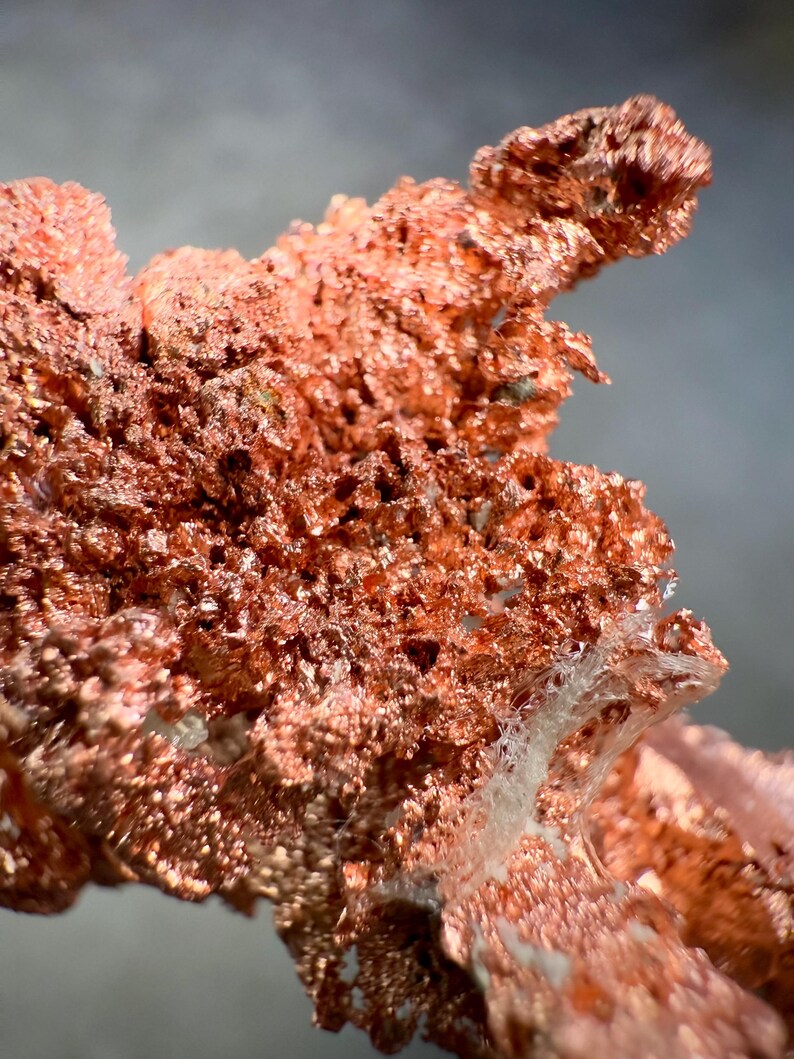 May include: Close-up of a raw copper specimen. The copper has a rough, textured surface with a reddish-brown color. The specimen has a crystalline structure with a porous appearance. The background is a soft gray.