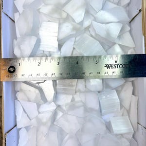 Pu&ograve; includere: Una scatola piena di pezzi di cristallo di selenite bianco traslucido extra piccoli. Un righello mostra che i pezzi sono lunghi circa 2,5-15 cm. Il testo "Extra Small" &egrave; visibile.