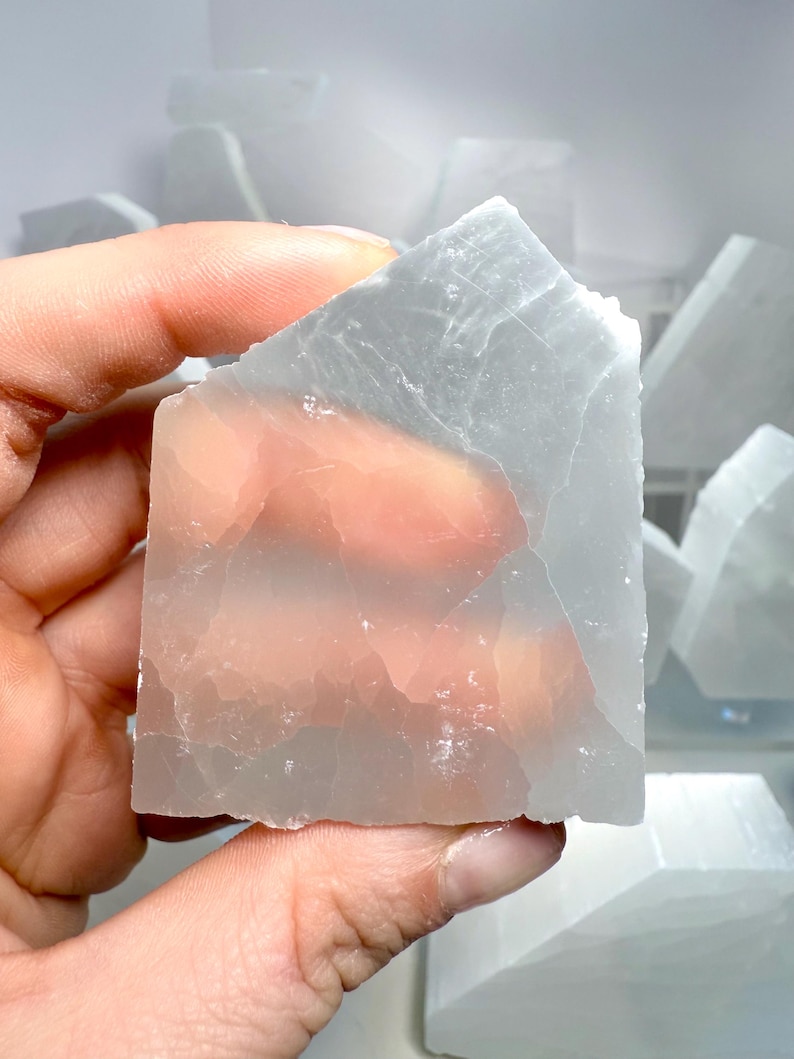 Pu&ograve; includere: Un cristallo di selenite traslucido, grigio chiaro, tenuto in mano. Il cristallo ha una consistenza ruvida e naturale con striature interne visibili. Altri pezzi di selenite sono visibili sullo sfondo.