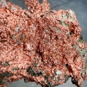 May include: A close-up of a raw copper specimen. The copper has a rough, textured surface with a vibrant reddish-brown color. Patches of green and dark gray minerals are visible throughout the specimen, adding contrast and detail to the natural formation.