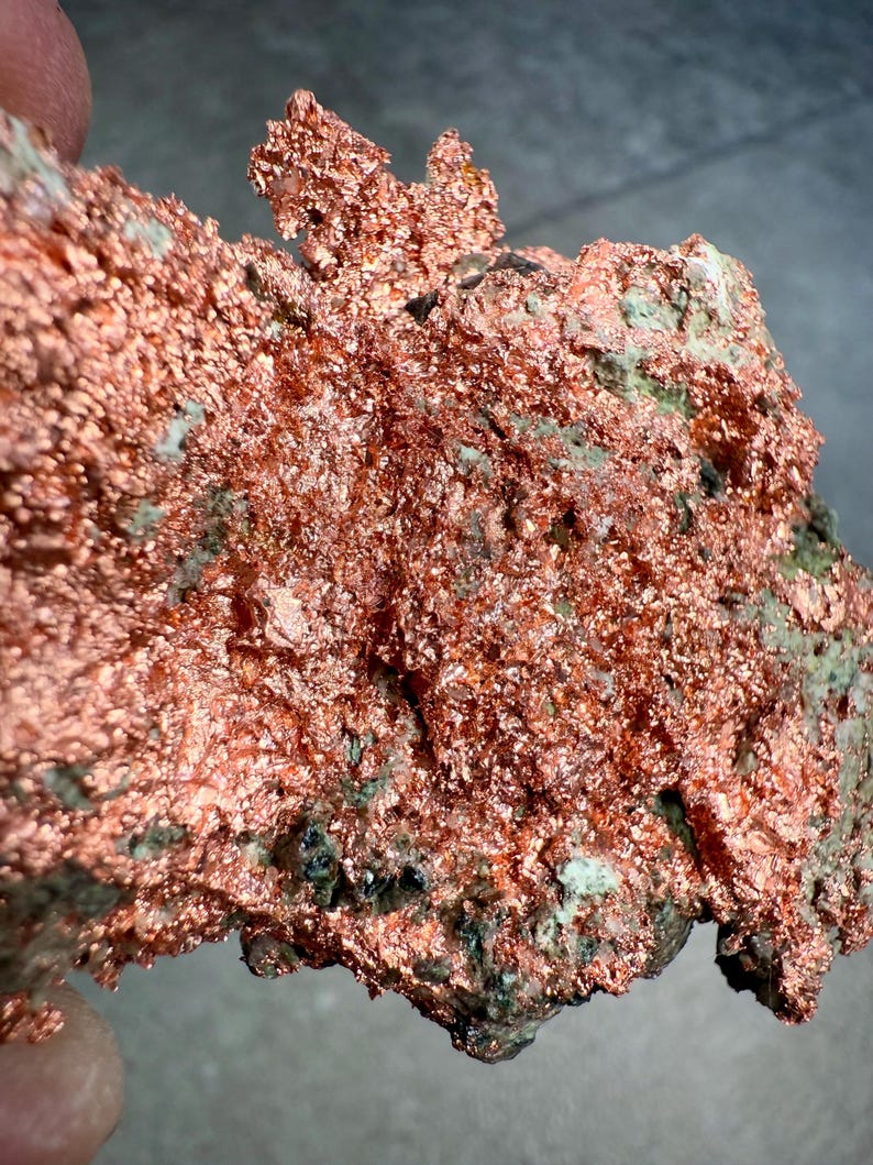 May include: A close-up of a raw copper specimen. The copper has a rough, textured surface with a vibrant reddish-brown color. Patches of green and dark gray minerals are visible throughout the specimen, adding contrast and detail to the natural formation.