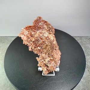 May include: A rough, reddish-brown mineral specimen, possibly a copper ore, is displayed on a black rotating platform. The mineral has a textured surface and is supported by a clear acrylic stand. The background is a neutral gray.