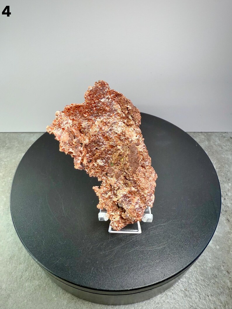 May include: A rough, reddish-brown mineral specimen, possibly a copper ore, is displayed on a black rotating platform. The mineral has a textured surface and is supported by a clear acrylic stand. The background is a neutral gray.