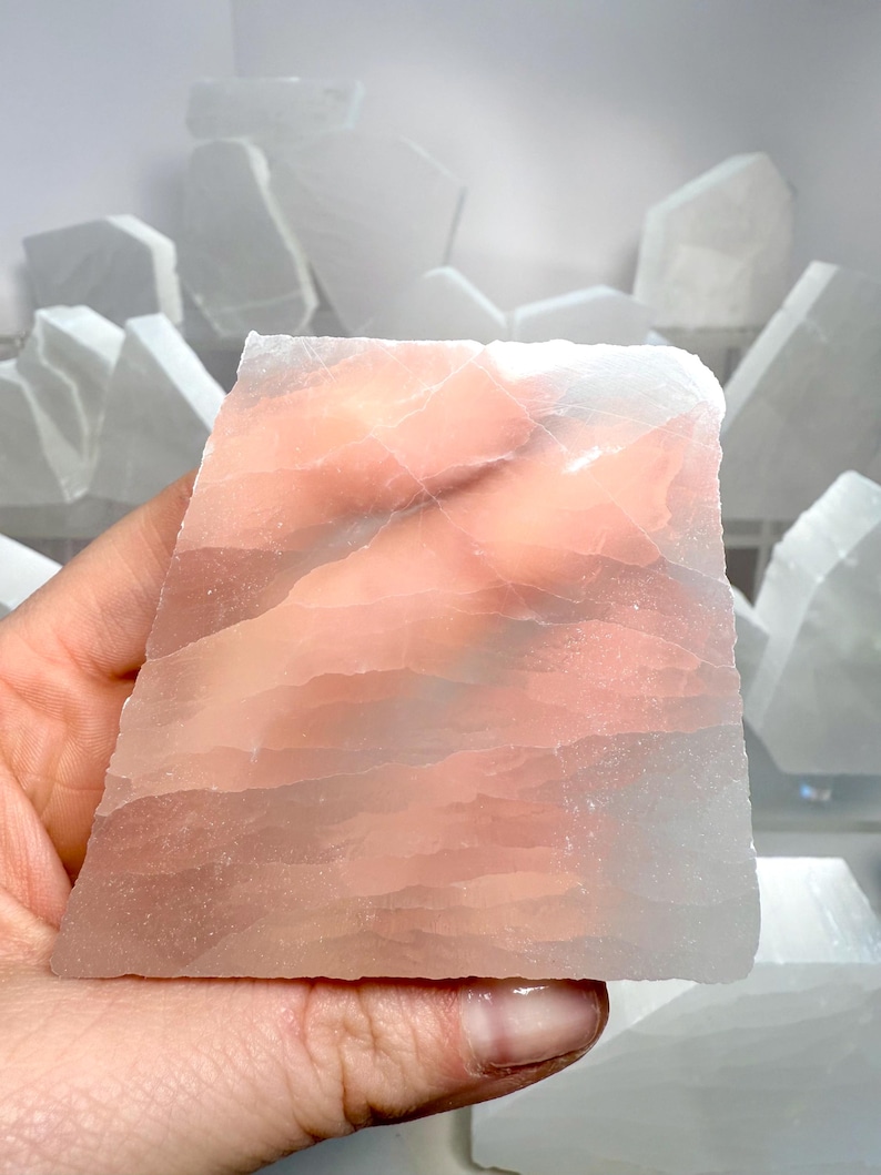 Pu&ograve; includere: Una mano che tiene un cristallo di selenite rettangolare traslucido con strati di tonalit&agrave; rosa e bianche. Altri pezzi di selenite sono visibili sullo sfondo, suggerendo una collezione di cristalli naturali.