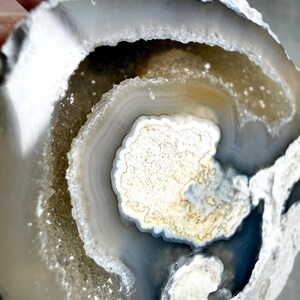 May include: A circular agate geode slice featuring concentric bands of white, gray, and translucent tan. The center contains a textured, crystalline cavity with sparkling druzy formations surrounded by smooth, layered mineral rings.