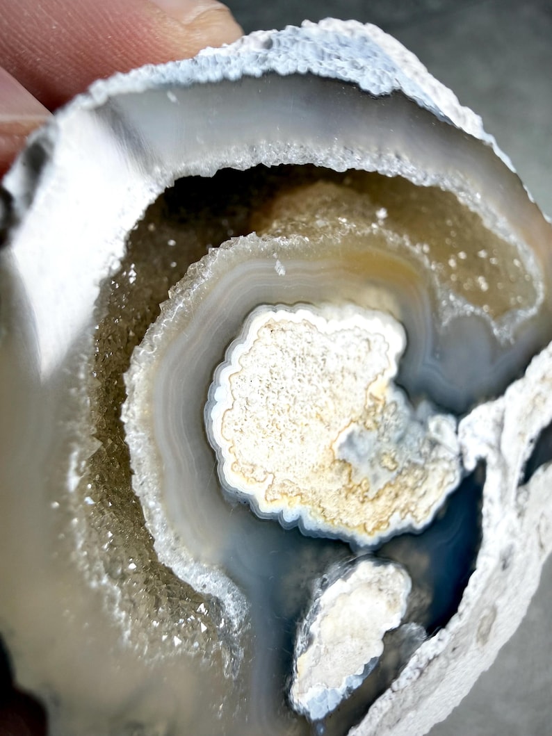 May include: A circular agate geode slice featuring concentric bands of white, gray, and translucent tan. The center contains a textured, crystalline cavity with sparkling druzy formations surrounded by smooth, layered mineral rings.