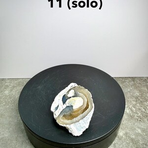May include: A geode half with a white, blue, and brown color scheme. The geode is on a black circular stand. The text "11 (solo)" is above the geode.