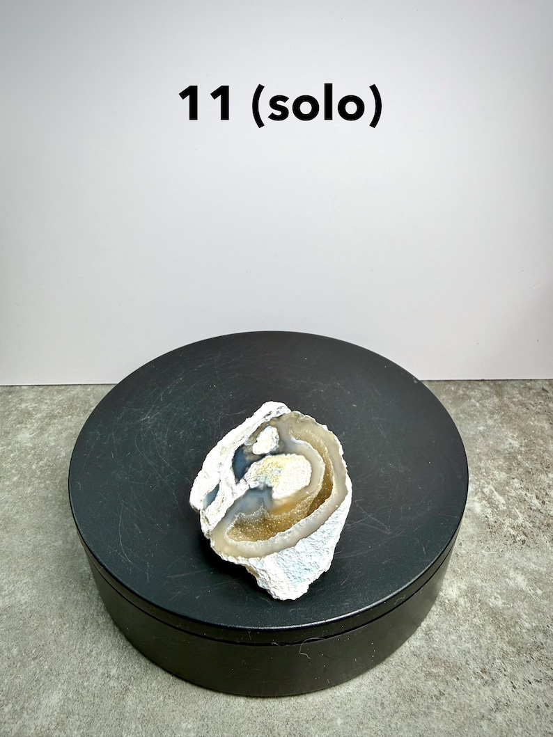 May include: A geode half with a white, blue, and brown color scheme. The geode is on a black circular stand. The text "11 (solo)" is above the geode.