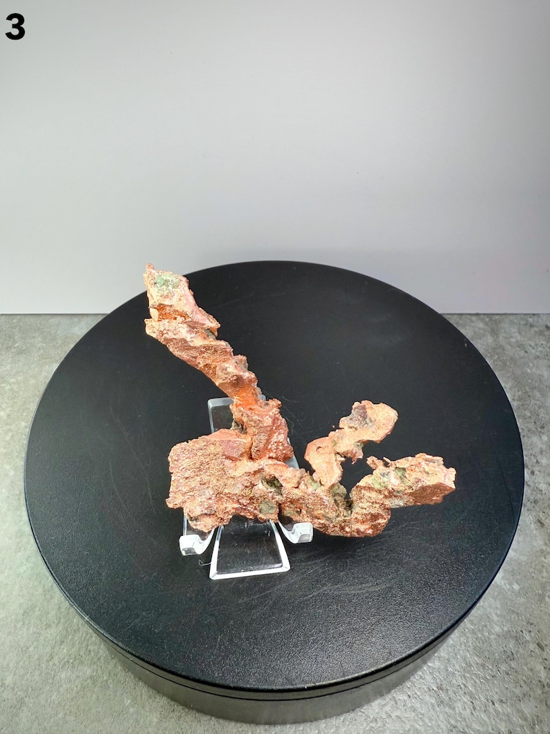 May include: A natural copper specimen with a rough, textured surface and a reddish-brown color. The copper is displayed on a clear acrylic stand, set against a black circular base. The number "3" is in the top left corner.
