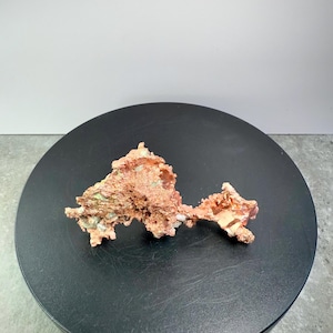 May include: A raw copper specimen with a rough, textured surface. The copper has a reddish-brown color with hints of green and white. The mineral is displayed on a black, circular platform.