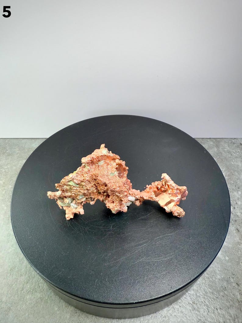 May include: A raw copper specimen with a rough, textured surface. The copper has a reddish-brown color with hints of green and white. The mineral is displayed on a black, circular platform.
