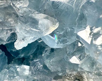 May include: Close-up of a celestite crystal cluster. The crystal formation features translucent, light blue, and clear crystalline structures. The edges of the crystals are sharp and angular, with a rough, textured base. The overall appearance is a natural, geological specimen.