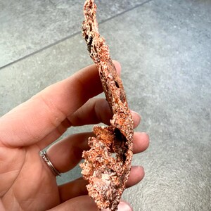 May include: A piece of raw copper ore, held in a hand. The copper has a rough, textured surface with a reddish-brown color. The ore has an irregular shape with holes and protrusions, suggesting a natural formation.