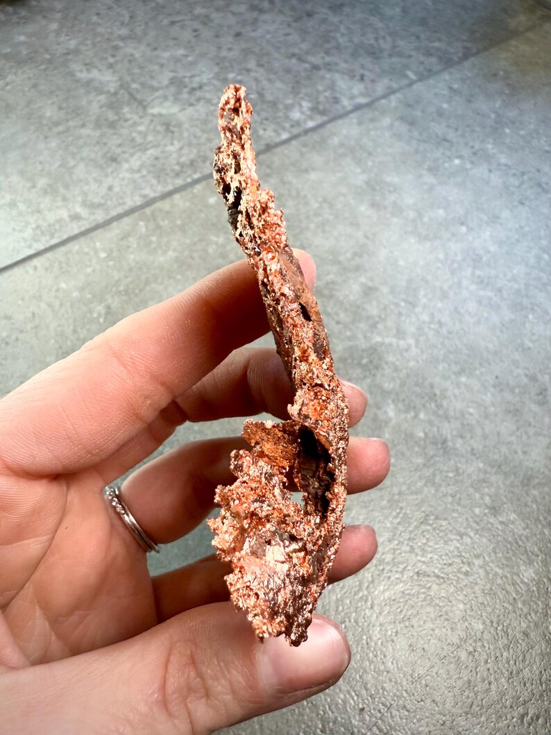 May include: A piece of raw copper ore, held in a hand. The copper has a rough, textured surface with a reddish-brown color. The ore has an irregular shape with holes and protrusions, suggesting a natural formation.