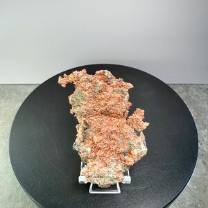 May include: A raw copper specimen with a rough, textured surface. The copper has a reddish-brown color with patches of green and is displayed on a white stand. The specimen is set against a black circular base and a white background.