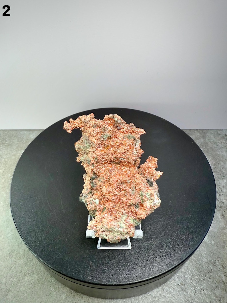 May include: A raw copper specimen with a rough, textured surface. The copper has a reddish-brown color with patches of green and is displayed on a white stand. The specimen is set against a black circular base and a white background.