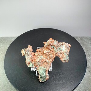 May include: A natural copper specimen with a rough, textured surface. The mineral displays a mix of reddish-brown and green hues, resting on a clear stand. The specimen is displayed on a black circular platform against a neutral background.