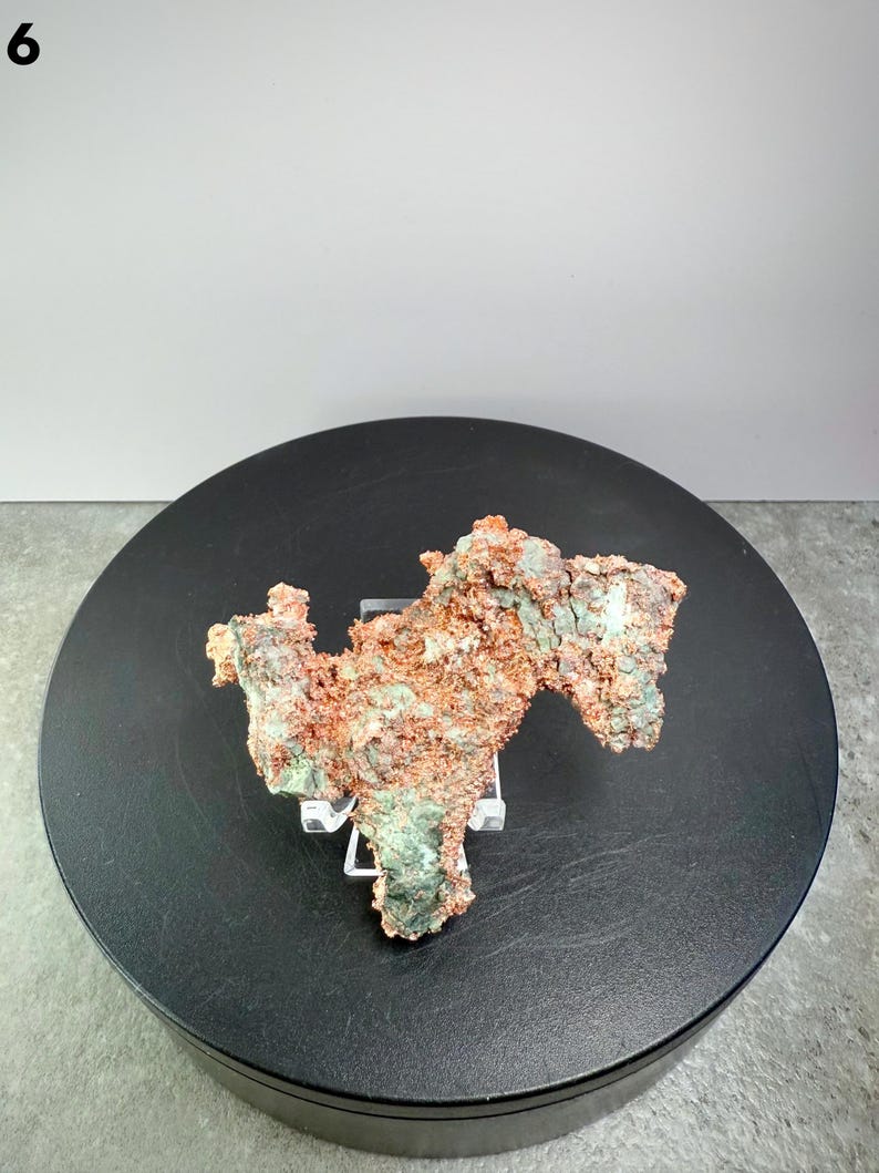 May include: A natural copper specimen with a rough, textured surface. The mineral displays a mix of reddish-brown and green hues, resting on a clear stand. The specimen is displayed on a black circular platform against a neutral background.
