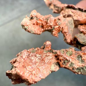 May include: Close-up of raw copper ore with a rough, textured surface. The copper has a reddish-brown color with patches of green and gray oxidation. The irregular shapes suggest a natural, unrefined state.