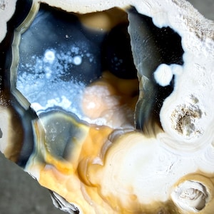 May include: A close-up of a geode with a hollow center. The geode features layers of white, brown, yellow, and blue colors. The interior has a dark blue and black hue with white speckles. The outer edges are a creamy white color.