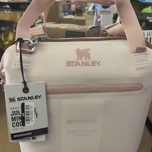 May include: A light pink Stanley cooler bag with a matching handle and shoulder strap. The bag has a zippered top and a tag with product information. The Stanley logo is visible on the front.