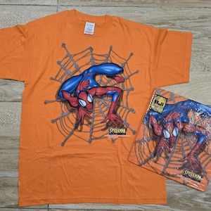 Puede incluir: Camiseta naranja con un dise&ntilde;o de Spider-Man dentro de una telara&ntilde;a. El superh&eacute;roe se muestra con su traje rojo y azul cl&aacute;sico. Tambi&eacute;n se ve una versi&oacute;n empaquetada del mismo dise&ntilde;o.