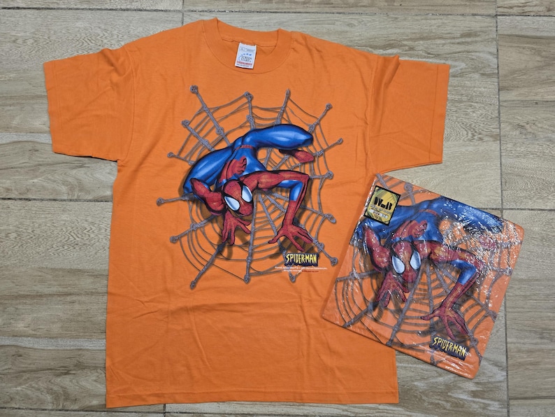 Puede incluir: Camiseta naranja con un dise&ntilde;o de Spider-Man dentro de una telara&ntilde;a. El superh&eacute;roe se muestra con su traje rojo y azul cl&aacute;sico. Tambi&eacute;n se ve una versi&oacute;n empaquetada del mismo dise&ntilde;o.