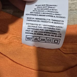 Puede incluir: Primer plano de una camiseta naranja con una etiqueta de cuidado adjunta. La etiqueta incluye instrucciones de lavado y texto en varios idiomas, incluidos ingl&eacute;s, alem&aacute;n y franc&eacute;s. La etiqueta tambi&eacute;n indica que la camisa fue fabricada en Marruecos.