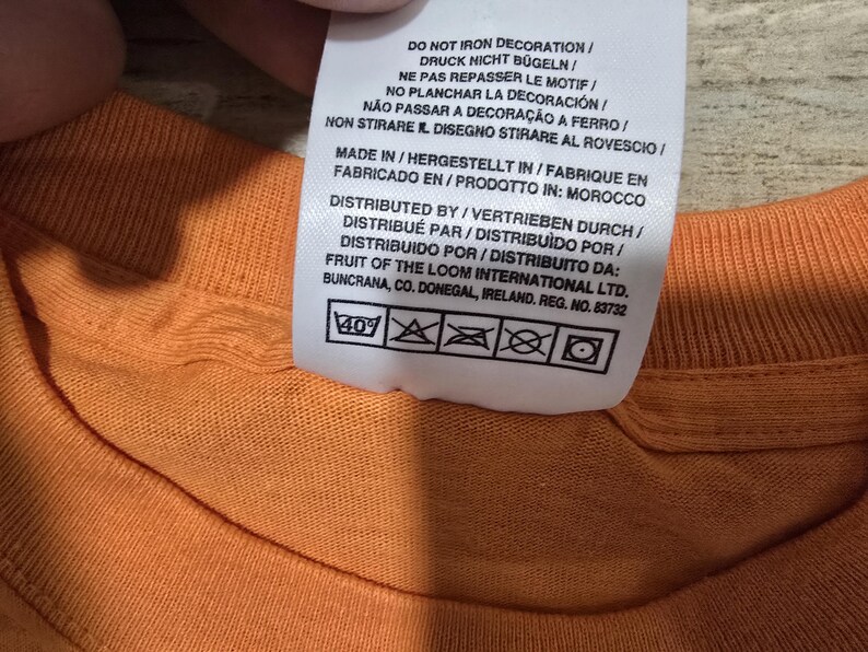 Puede incluir: Primer plano de una camiseta naranja con una etiqueta de cuidado adjunta. La etiqueta incluye instrucciones de lavado y texto en varios idiomas, incluidos ingl&eacute;s, alem&aacute;n y franc&eacute;s. La etiqueta tambi&eacute;n indica que la camisa fue fabricada en Marruecos.