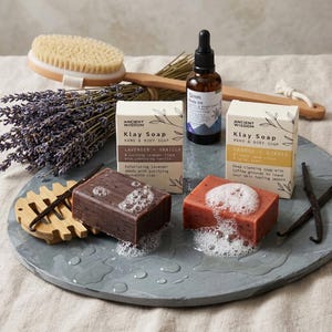 Pode incluir: Dois sabonetes retangulares Klay Soap, lavanda e baunilha e laranja e gengibre, com espuma. Inclui um frasco de óleo corporal, uma escova de madeira, lavanda seca e favas de baunilha numa bandeja redonda de pedra cinzenta.