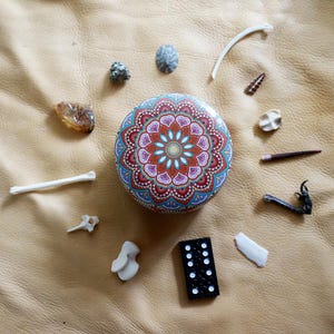 May include: A round tin featuring a colorful, symmetrical mandala pattern in shades of orange, teal, and white, surrounded by a collection of small natural objects including various seashells, small animal bones, a piece of amber, and a black domino.
