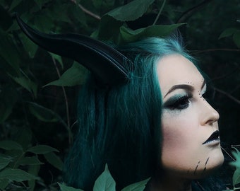 May include: A person with teal hair and dark makeup, wearing a horned headpiece and ornate jewelry, poses amidst lush green foliage. The image has a dark, gothic aesthetic, with a focus on fantasy and alternative fashion.