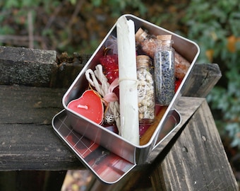May include: A silver metal box filled with various items, including a rolled-up parchment, small glass bottles with cork stoppers, a red heart-shaped candle, and bundles of twine. The box sits on a wooden surface.