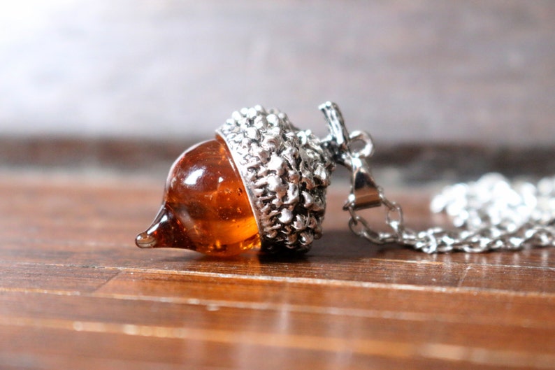 May include: A silver acorn pendant necklace with a clear amber glass acorn. The necklace is on a silver chain.