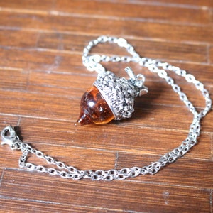 May include: A silver chain necklace with an acorn pendant. The acorn is made of glass and has a brown and amber color.