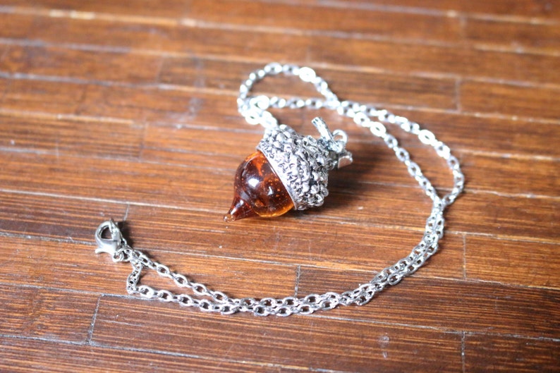 May include: A silver chain necklace with an acorn pendant. The acorn is made of glass and has a brown and amber color.