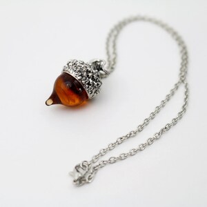 May include: A silver chain necklace with an acorn pendant. The acorn is made of glass and has a brown color.