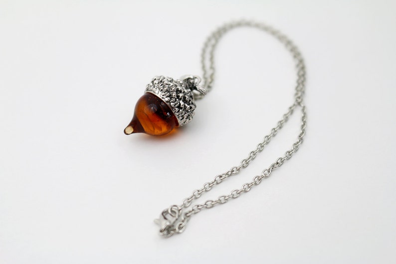 May include: A silver chain necklace with an acorn pendant. The acorn is made of glass and has a brown color.