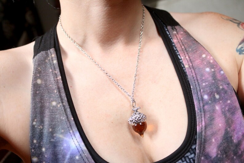 May include: A silver chain necklace with an acorn pendant. The acorn is made of glass and has a brown color.