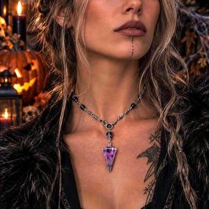 Peut inclure: Un collier argenté doté d'un pendentif en cristal violet translucide en forme de pointe. La chaîne est ornée de petites perles violet foncé et se connecte au pendentif par une bélière en métal. Le collier repose sur le cou, mettant en valeur le cristal.