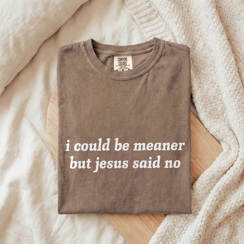 May include: A brown Comfort Colors t-shirt with the text "i could be meaner but jesus said no" in white. The shirt is folded and placed on a wooden board, with a cream-colored blanket in the background.