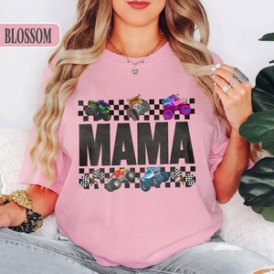 May include: Pink t-shirt with the word "MAMA" in large black letters. Above the word are six colorful monster trucks on a checkered background. The word "BLOSSOM" is in the upper left corner.