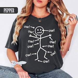 May include: Dark gray t-shirt with a white stick figure design. The stick figure has arrows pointing to it with the word "ow!" written next to each arrow. The word "PEPPER" is in a black rectangle in the upper left corner.
