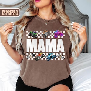May include: Espresso-colored t-shirt featuring the word "MAMA" in large white letters, with colorful monster truck graphics above and below. The design includes checkered flags and various monster truck designs in green, orange, pink, red, and blue.