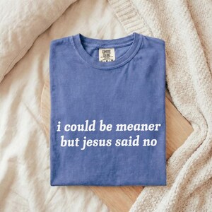 May include: A blue Comfort Colors t-shirt with the text "i could be meaner but jesus said no" in white. The shirt is folded on a wooden board, with a cream-colored blanket and pillow in the background.