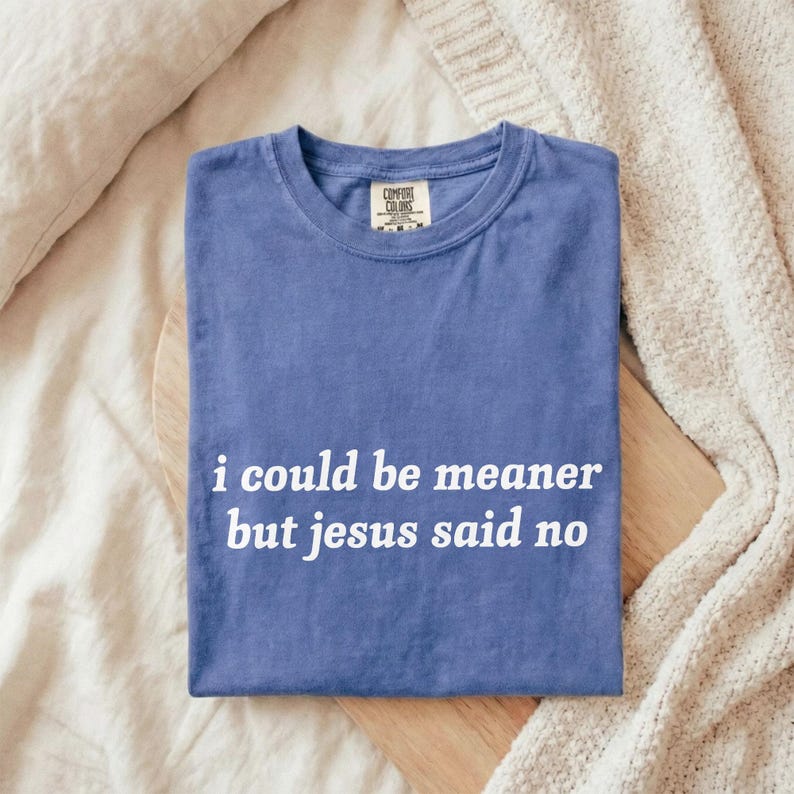 May include: A blue Comfort Colors t-shirt with the text "i could be meaner but jesus said no" in white. The shirt is folded on a wooden board, with a cream-colored blanket and pillow in the background.