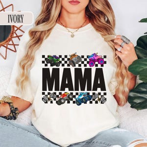 May include: Ivory-colored t-shirt featuring the word "MAMA" in large black letters. Above and below the word are checkered patterns with colorful monster truck graphics. The trucks are in various colors, including green, orange, red, and blue.