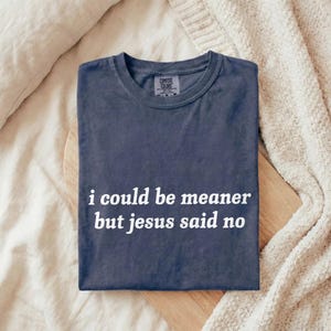 May include: A navy blue Comfort Colors t-shirt with the text "i could be meaner but jesus said no" in white. The shirt is folded on a wooden cutting board, with a cream-colored blanket and pillow in the background.