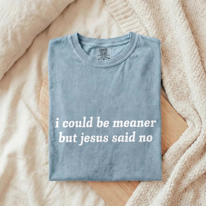 May include: A light blue t-shirt with the text "i could be meaner but jesus said no" in white. The shirt is folded and rests on a wooden surface, with a cream-colored blanket in the background.