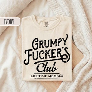 May include: Ivory-colored t-shirt with the text "Grumpy Fucker's Club" in a bold, distressed black font. The words "Lifetime Member" are printed below the club name. The shirt is laid flat on a wooden surface.